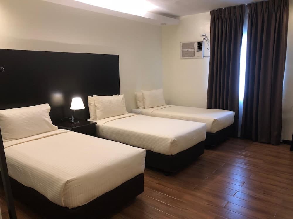 A cozy hotel room with three neatly arranged single beds, wooden flooring, soft lighting from a bedside lamp, and a window with dark curtains, ideal for budget travelers seeking comfort and simplicity.