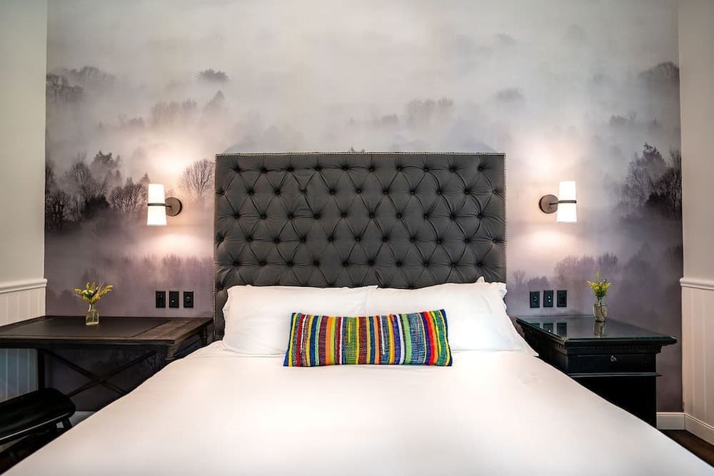 A cozy hotel room features a plush bed with a tufted headboard against a misty forest wallpaper, flanked by modern bedside tables with lamps and small vases, ideal for travelers seeking comfort and style.