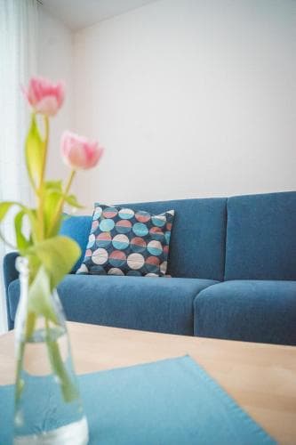A cozy living room scene featuring a blue sofa adorned with a colorful pillow, a wooden table with a vase of pink tulips, evoking a serene and inviting atmosphere ideal for a relaxing getaway or home stay.
