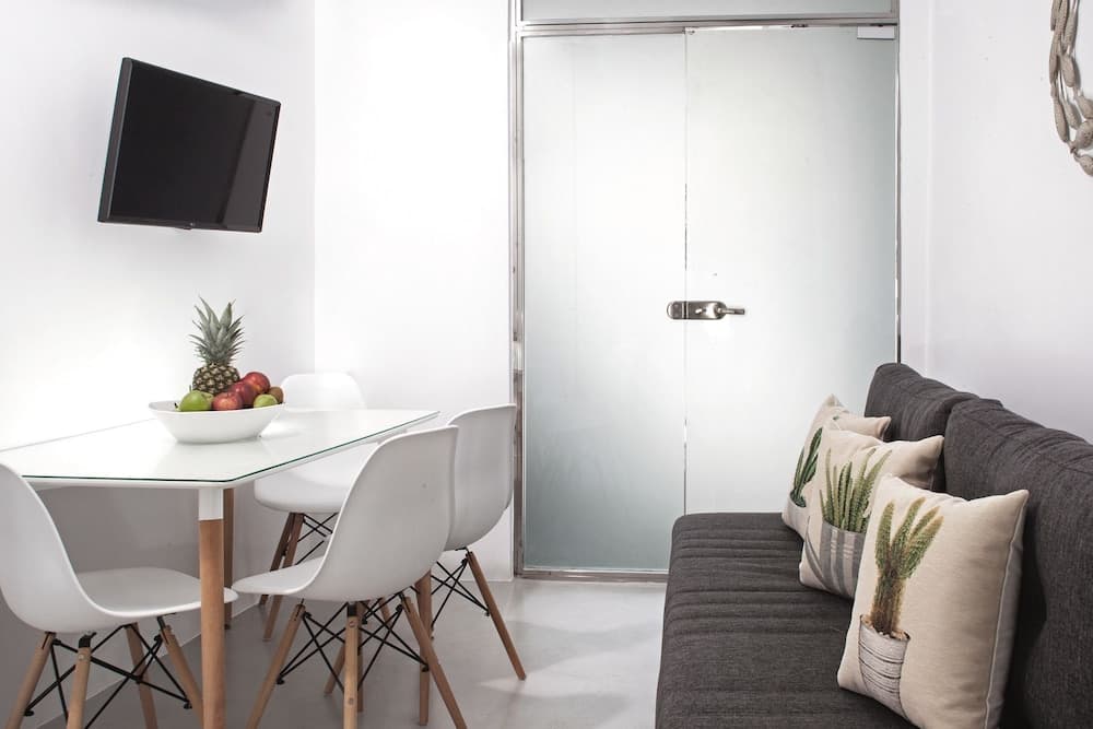 A cozy modern apartment dining area features a sleek glass table with fruit, white chairs, a gray sofa adorned with cactus-patterned pillows, and a minimalist vibe ideal for short-term rentals or vacation stays.