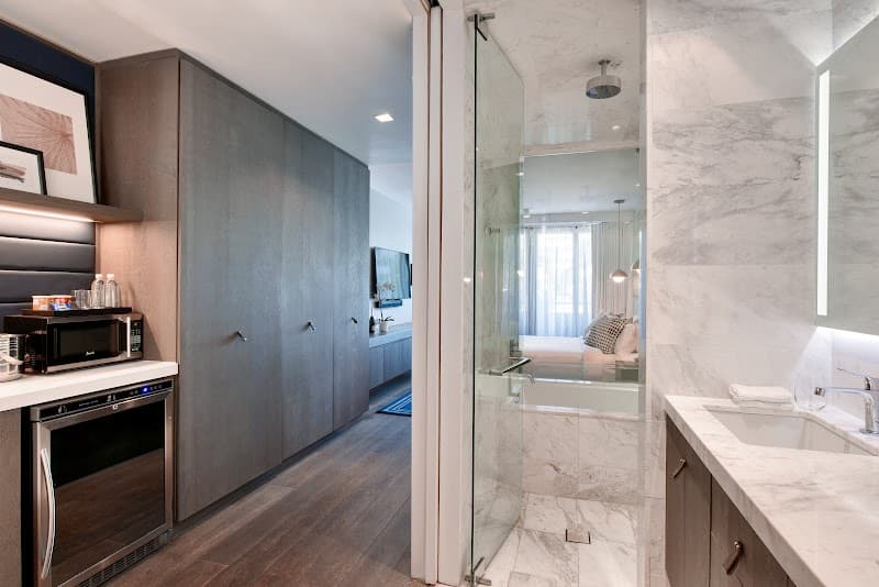 A luxurious hotel suite at Kaskades Hotel South Beach features a sleek kitchenette with modern appliances, a spacious marble bathroom with a glass-enclosed shower, and a cozy bedroom in the background, ideal for travelers seeking comfort and style in Miami Beach.