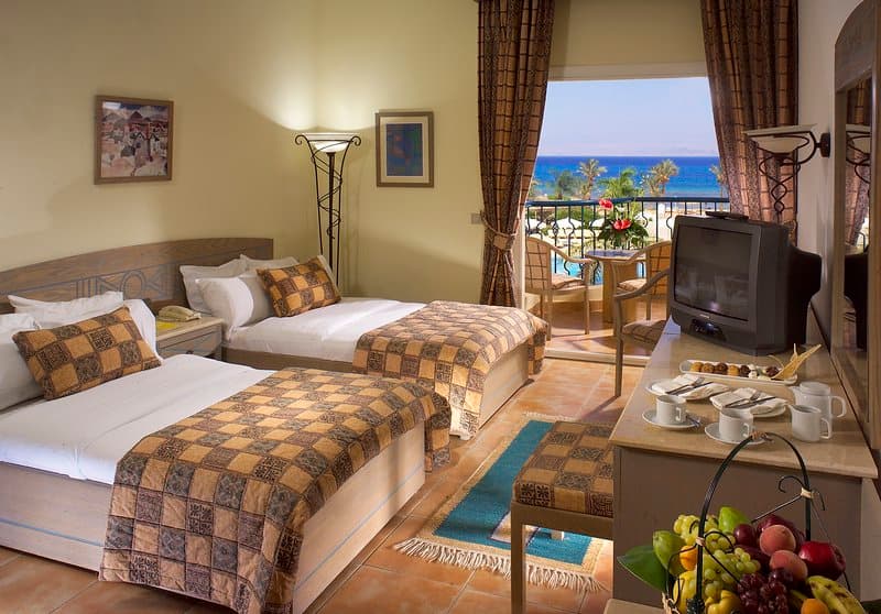 A cozy hotel room with twin beds adorned in checkered throws overlooks the serene ocean through a balcony, perfect for a relaxing seaside getaway.