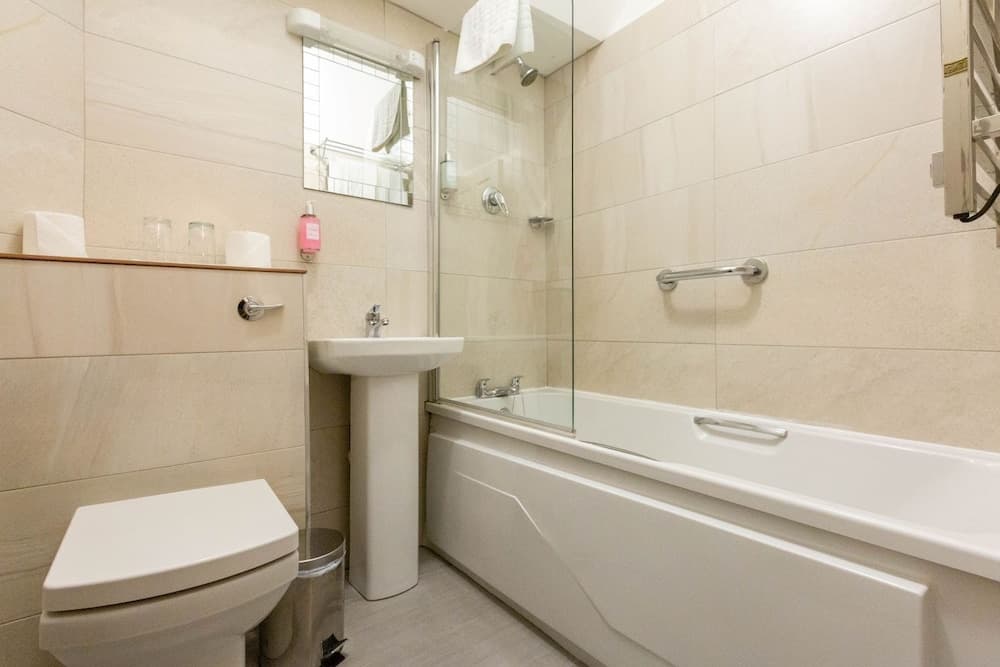 A clean, modern bathroom with beige tiles, a bathtub with a glass shower screen, a pedestal sink, and neatly arranged toiletries, ideal for travelers seeking comfort and hygiene in their accommodations.