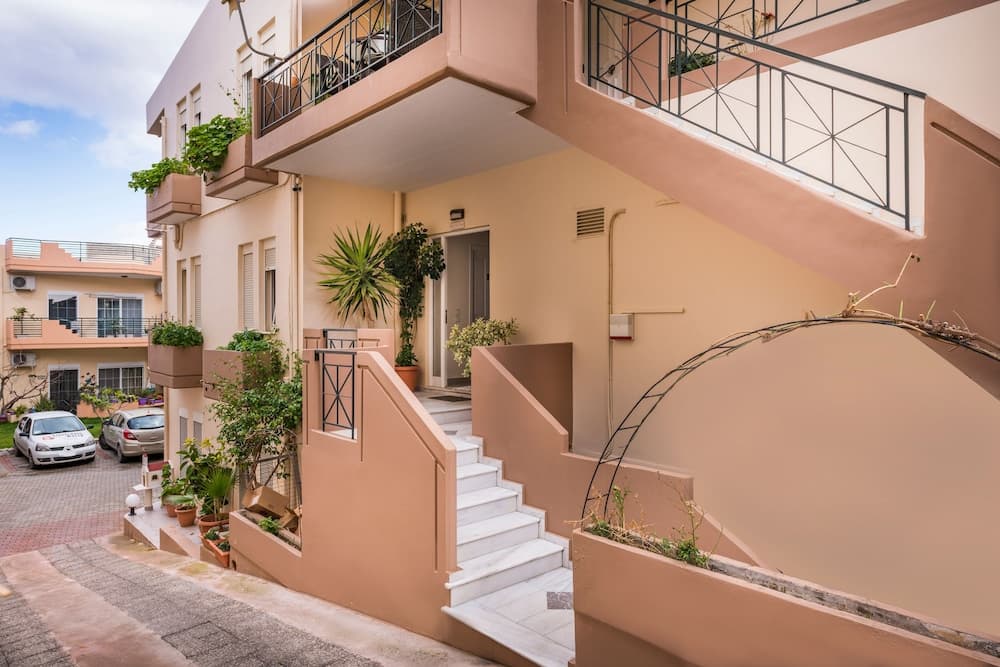 A charming, sunlit Mediterranean apartment entrance with terracotta walls, lush greenery, and a welcoming staircase invites travelers seeking cozy, picturesque accommodations.