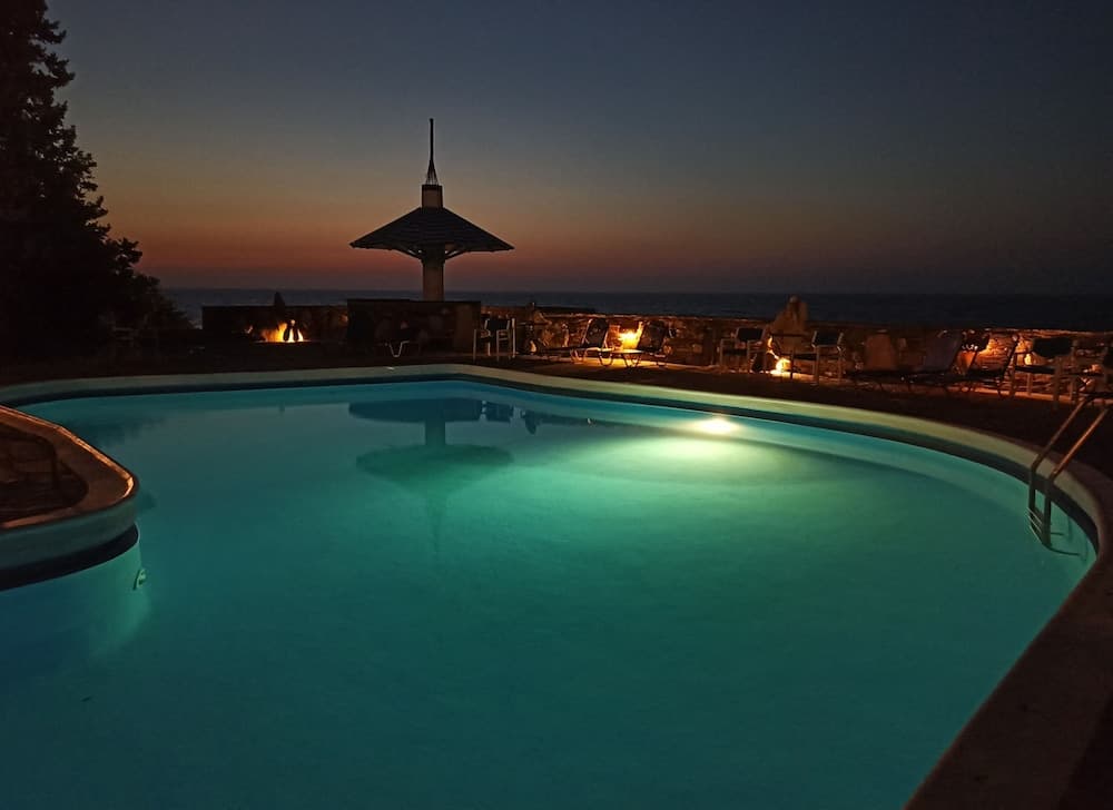 A serene evening at an oceanside resort features a glowing pool, cozy lounge chairs, and a pagoda-style structure under a twilight sky, perfect for a relaxing getaway.