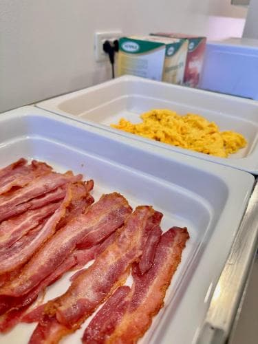A cozy hotel breakfast buffet scene featuring crispy bacon and scrambled eggs in white trays, ideal for travelers seeking a hearty morning meal.