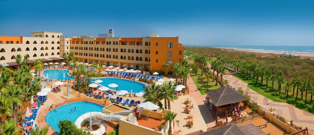 A vibrant beachside resort features expansive pools, palm-lined pathways, and colorful lounge areas under clear skies, ideal for family vacations seeking relaxation and fun.