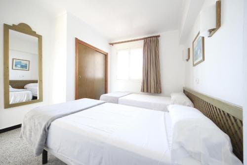 A cozy hotel room with white walls, a neatly made bed, a mirror reflecting the space, and soft curtains by the window, ideal for travelers seeking comfort and simplicity.
