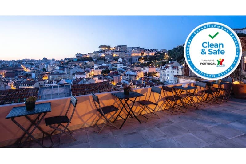 A serene rooftop terrace at Lisbon Hostel Costa do Castelo offers panoramic views of Lisbon's illuminated skyline, featuring cozy seating arrangements and a "Clean & Safe" certification badge, ideal for travelers seeking a tranquil evening spot in the city.