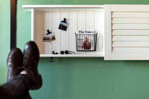 A cozy indoor scene features a person relaxing with feet up against a green wall, near a white shelf holding personal items like photos and a magazine titled "The Tides," ideal for a travel blog post about home relaxation spots.