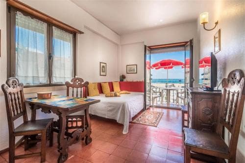 A cozy beachfront apartment features a vibrant interior with tiled floors, wooden furniture, and an inviting balcony view of the ocean under red umbrellas, perfect for a relaxing seaside getaway.