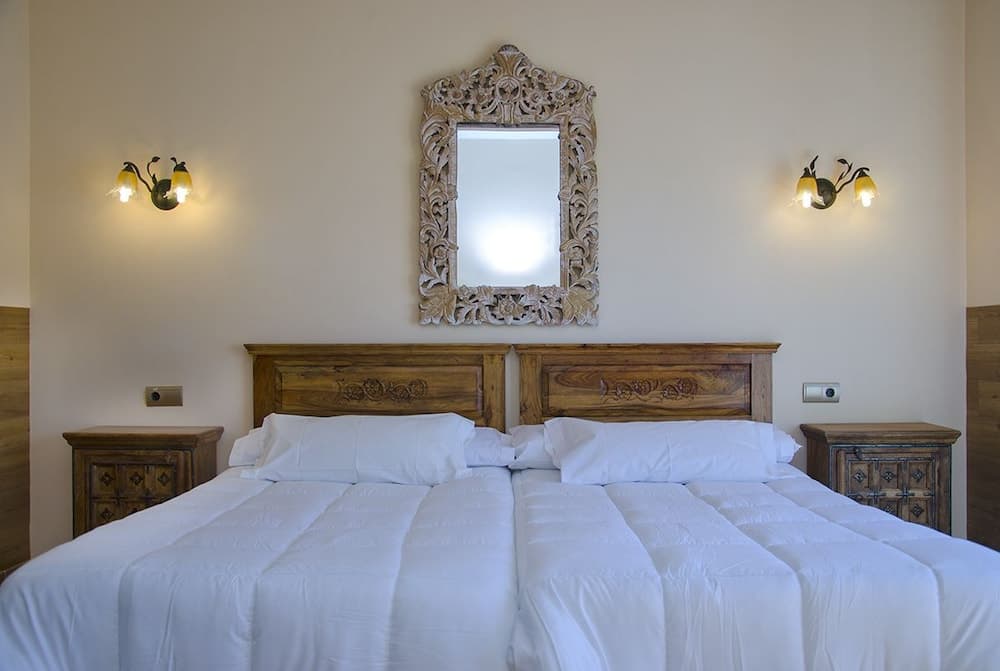 A cozy hotel bedroom features a neatly made bed with white linens, flanked by wooden nightstands and illuminated by wall-mounted lamps, creating a warm and inviting atmosphere ideal for travelers seeking comfort and relaxation.