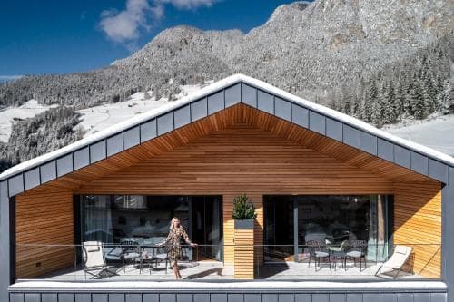 A modern wooden chalet with large glass windows overlooks a serene snowy mountain landscape, offering an ideal winter retreat for travelers seeking luxury and tranquility.