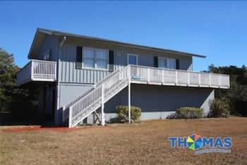 A serene coastal vacation home with a spacious deck and staircase stands amidst dry grass under a clear blue sky, ideal for family retreats seeking relaxation and beach proximity.