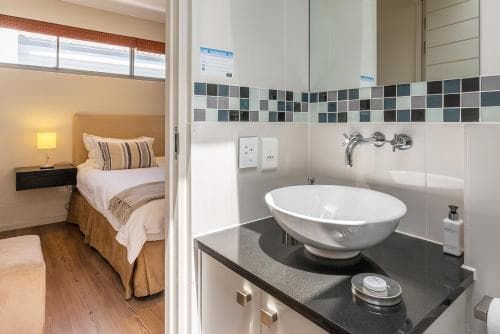 A cozy hotel room with a neatly made bed and bedside lamp is partially visible through an open door leading to a modern bathroom featuring a sleek sink, tiled walls, and a mirror, ideal for travelers seeking comfort and convenience.