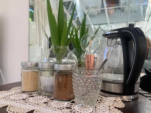 A cozy kitchen corner features a lace-covered table adorned with jars of spices, a glass cup, a modern electric kettle, and a vibrant potted plant, creating an inviting atmosphere perfect for travelers seeking a homey breakfast spot.