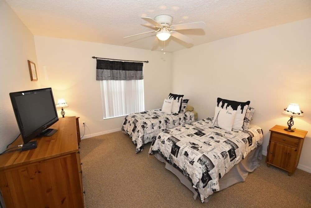 A cozy hotel room features two neatly made beds with Paris-themed bedding, warm lighting from bedside lamps, a wooden dresser with a TV, and a ceiling fan, ideal for travelers seeking comfort and relaxation.