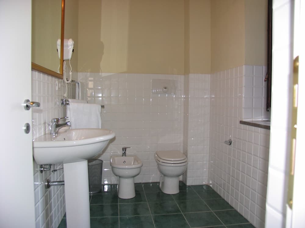 A clean and functional bathroom with white tiled walls, green floor tiles, a sink, bidet, toilet, wall-mounted hairdryer, and mirror, ideal for travelers seeking basic amenities in a compact space.
