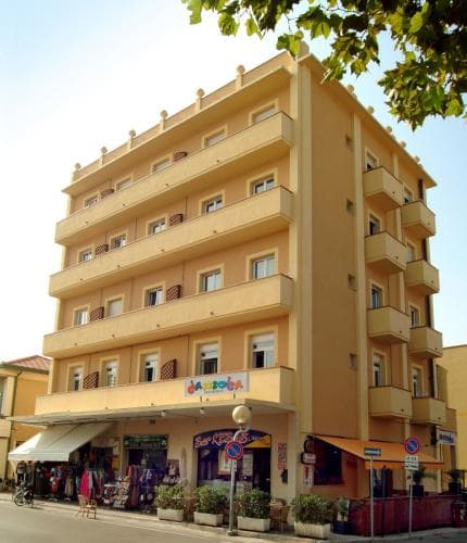 A sunny day highlights a vibrant yellow hotel with balconies, shops below, and lush greenery, ideal for travelers seeking a cozy urban retreat in a lively city setting.