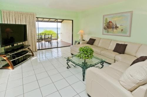 A cozy living room with white tiled floors, comfortable sofas, and a glass coffee table opens to a serene ocean view through sliding glass doors, ideal for a relaxing beachside vacation retreat.