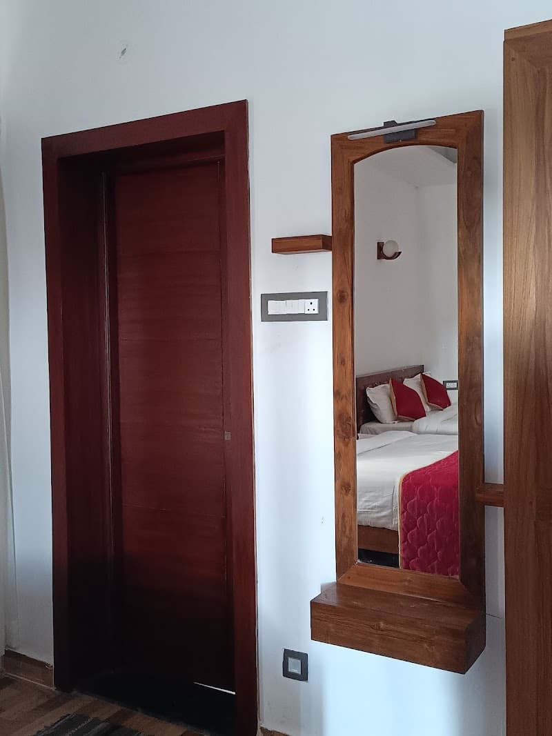 A cozy hotel room in Kodaikanal features a wooden-framed mirror reflecting a neatly made bed with red accents, creating a warm and inviting atmosphere ideal for travelers seeking comfort and relaxation.