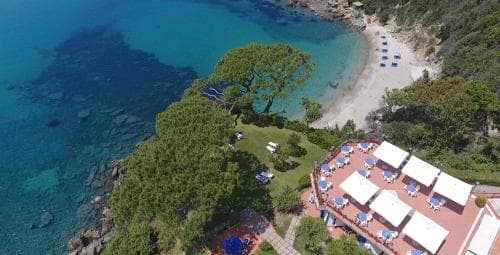 An aerial view captures a serene coastal resort with white umbrellas and lounge chairs nestled among lush greenery beside turquoise waters, ideal for travelers seeking relaxation and scenic beauty.