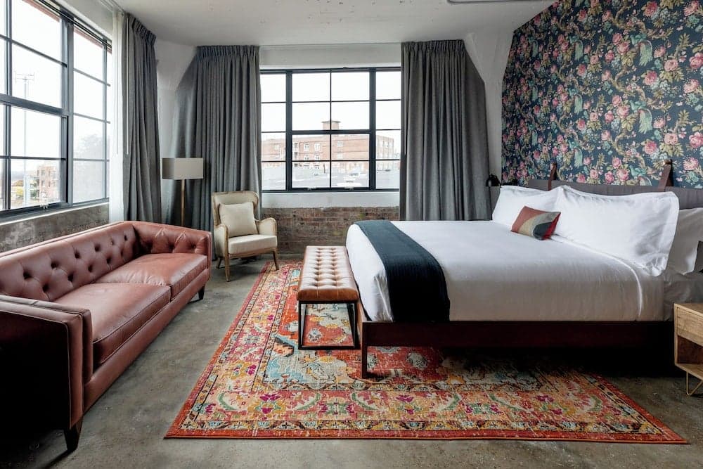 A cozy hotel room features an inviting bed beside a leather sofa, vibrant rug, and floral wallpaper, creating a stylish retreat perfect for travelers seeking comfort and charm.