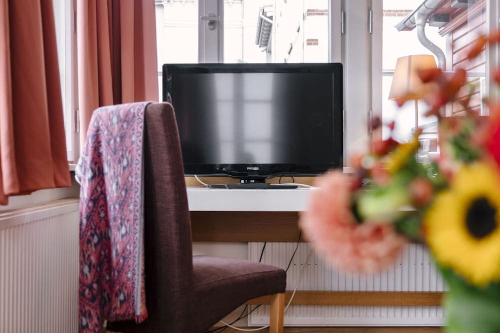 A cozy hotel room workspace features a desk with a flat-screen TV, a chair draped with a patterned scarf, warm-toned curtains, and vibrant flowers in the foreground, ideal for travelers seeking comfort and functionality.