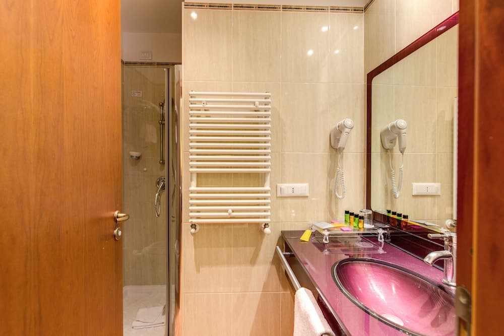 A cozy hotel bathroom features a sleek glass shower, a radiant towel warmer, a vibrant pink sink, and a mounted hairdryer, ideal for travelers seeking comfort and style.