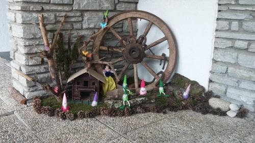 A whimsical garden scene featuring colorful gnomes, a rustic wagon wheel, and a miniature house nestled against a stone wall, perfect for a family-friendly outdoor adventure or a fairy tale-themed photo opportunity.