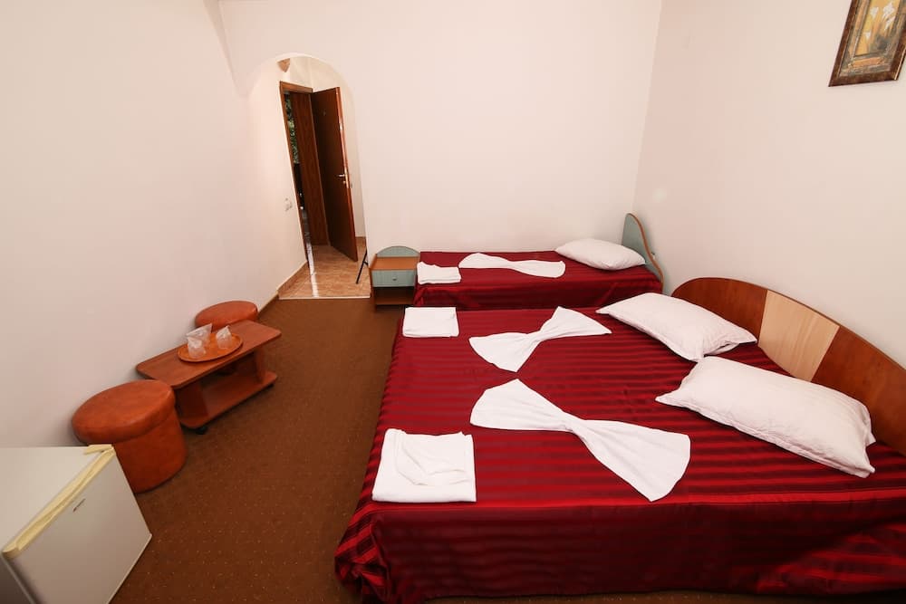 A cozy hotel room features two neatly made beds with red striped bedding, a small table with orange stools, and an inviting arched doorway leading to another space, perfect for travelers seeking comfort and simplicity.