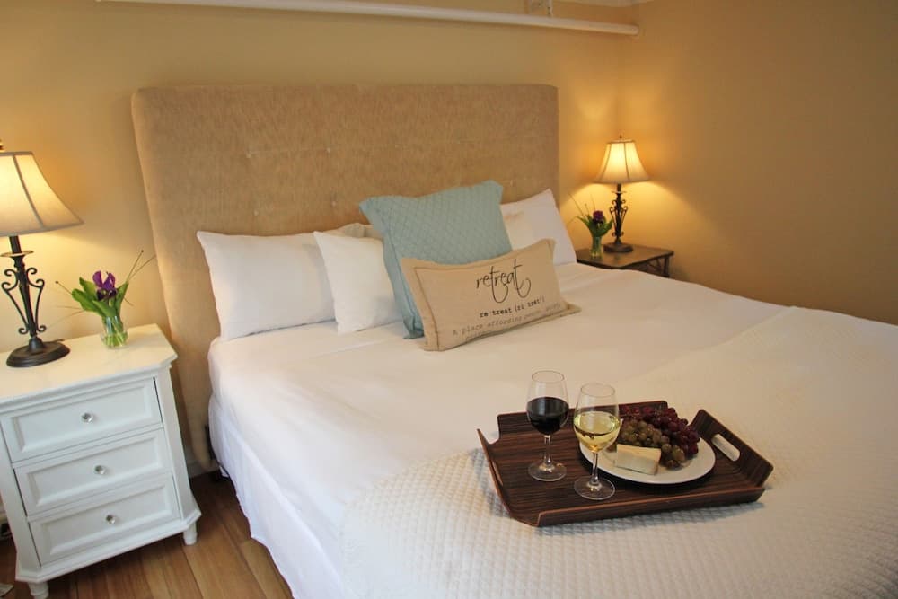 A cozy hotel room features a neatly made bed adorned with a "retreat" pillow, a tray of wine and cheese, soft lighting from bedside lamps, and a tranquil ambiance ideal for relaxation or romantic getaways.
