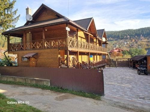 A cozy wooden lodge with balconies nestled in a serene mountain village, featuring a stone-paved driveway and surrounded by lush greenery, ideal for travelers seeking a tranquil retreat.
