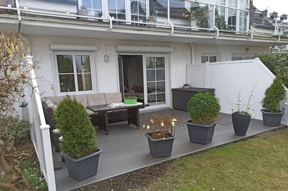 A cozy backyard patio features a comfortable seating area with a sofa and table, surrounded by potted plants and a white fence, ideal for relaxing or hosting small gatherings in a serene suburban setting.