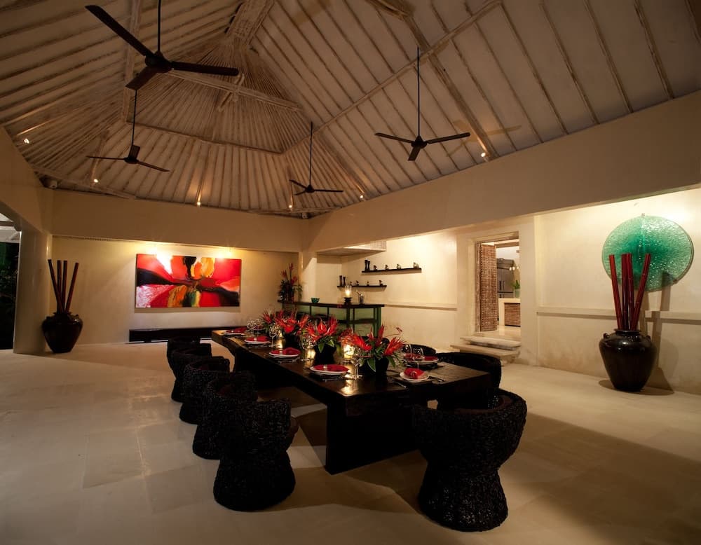 A warmly lit tropical dining area with a thatched roof, vibrant artwork, and elegant table setup creates an inviting ambiance ideal for luxury resort stays.