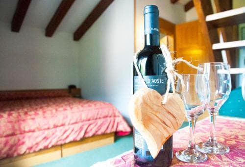 A cozy bedroom with rustic wooden beams sets the stage for relaxation, featuring a bottle of Valle d'Aosta wine and two glasses on a vibrant tablecloth, perfect for a romantic getaway or wine-tasting retreat.