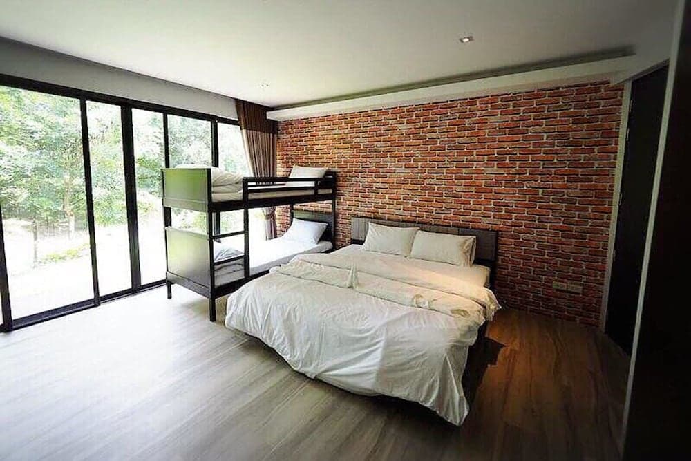 A cozy bedroom with a brick accent wall, bunk beds, and a large bed features a serene view of greenery through floor-to-ceiling windows, ideal for travelers seeking comfort in a modern lodge setting.