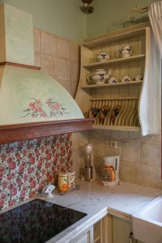 A cozy kitchen with floral wallpaper, a decorative range hood, and open shelving filled with ceramic dishes exudes a charming, rustic vibe ideal for travelers seeking a homely culinary experience.