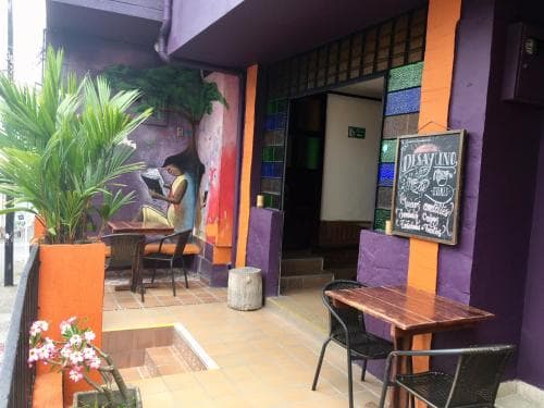 A cozy outdoor café with vibrant purple walls, colorful glass blocks, and potted plants creates an inviting atmosphere perfect for travelers seeking a relaxing spot to enjoy coffee or a meal.