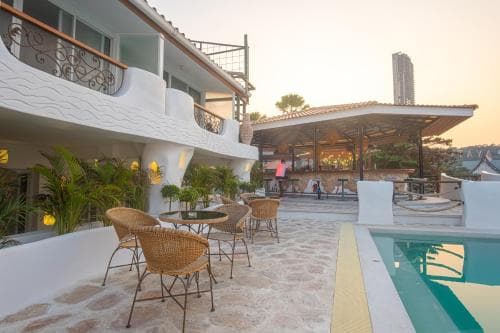 A serene tropical resort patio features a sparkling pool, wicker lounge chairs, lush greenery, and a modern building backdrop, ideal for a relaxing vacation getaway.