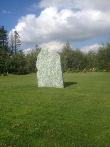 A serene park scene features a large textured stone sculpture standing prominently on lush green grass under a partly cloudy sky, ideal for nature lovers seeking tranquil outdoor art installations.