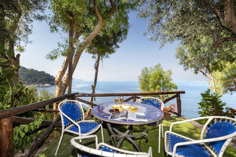 A serene coastal patio with blue cushioned chairs and a table set for breakfast under lush green trees, perfect for a tranquil seaside getaway.