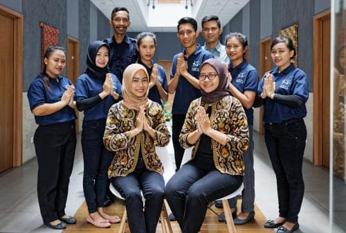 A welcoming group of staff in traditional attire greets visitors with warm smiles and hands pressed together in a serene indoor setting, ideal for showcasing hospitality services in cultural tourism.