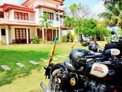 A serene tropical villa with lush greenery and a parked Royal Enfield motorcycle adorned with flags creates an inviting atmosphere for adventure travelers seeking a peaceful retreat.