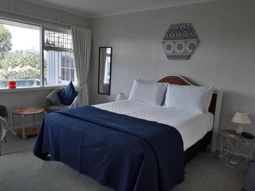 A cozy hotel room features a neatly made bed with blue bedding, a decorative wall hanging, and natural light streaming through large windows, creating a serene atmosphere ideal for travelers seeking relaxation.