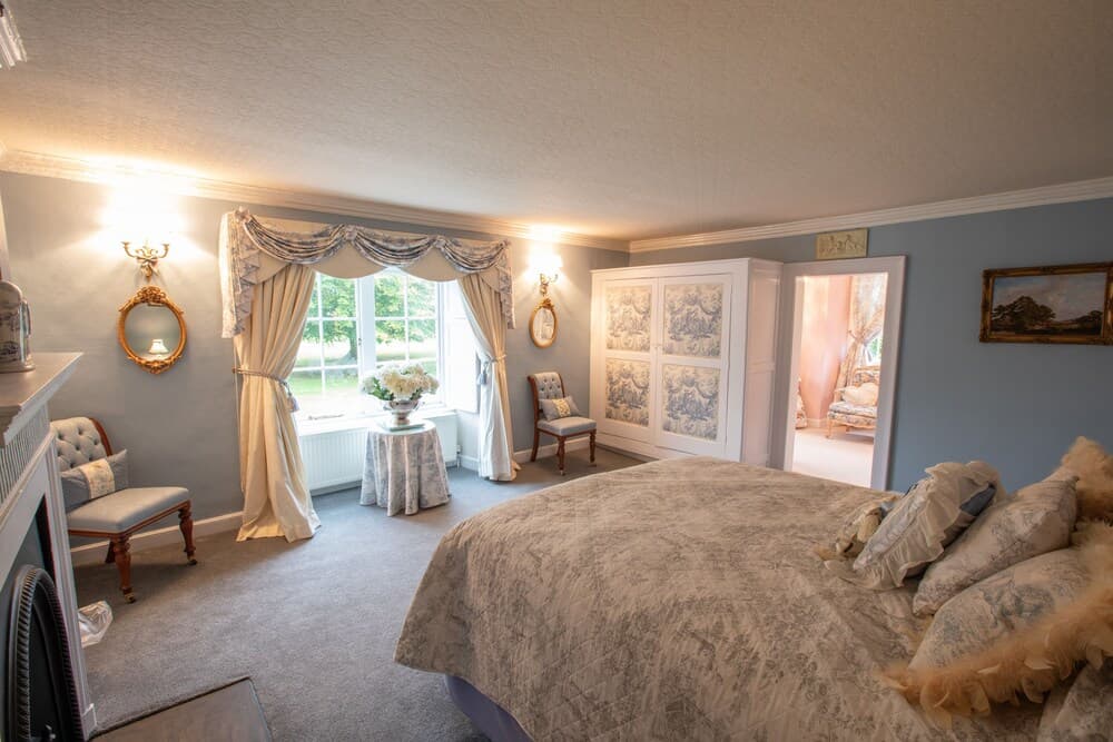A cozy, elegantly furnished bedroom with soft blue walls, ornate mirrors, and a large window overlooking greenery, ideal for a serene getaway or boutique hotel stay.