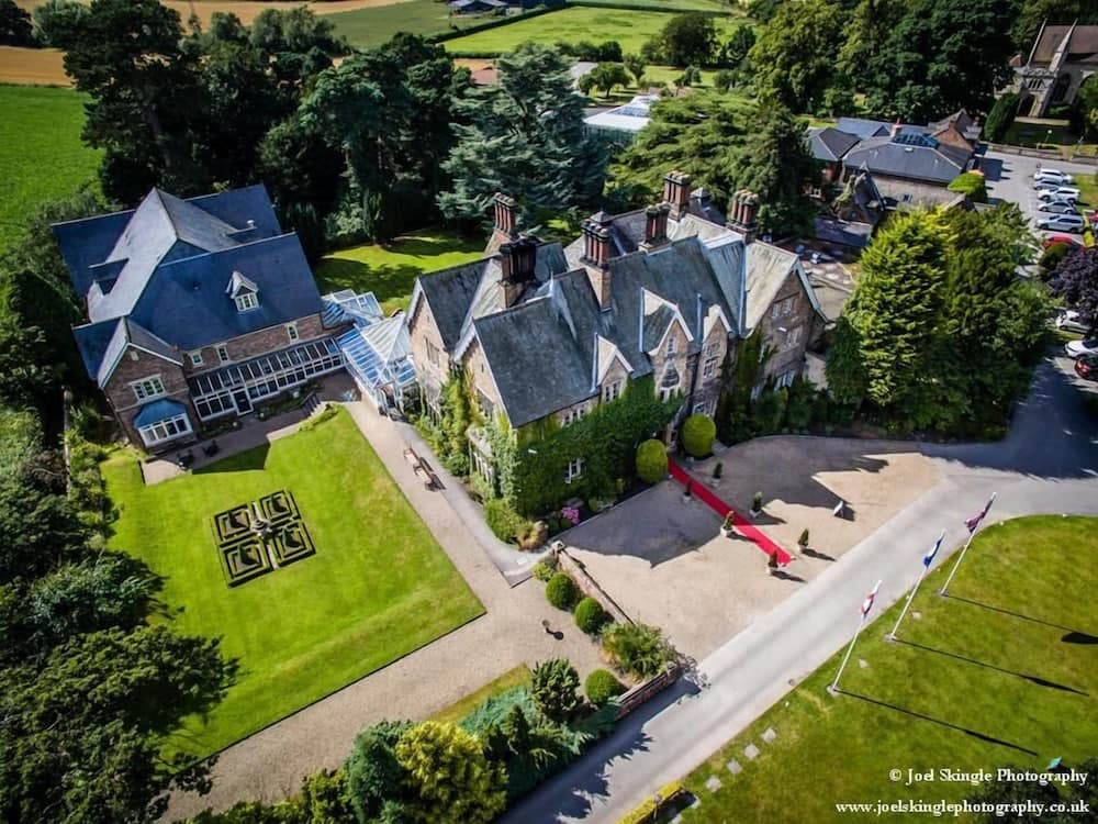 An aerial view of an elegant countryside estate with lush greenery, a manicured lawn featuring geometric patterns, and a grand driveway leading to a historic building, ideal for luxury retreats and wedding venues.