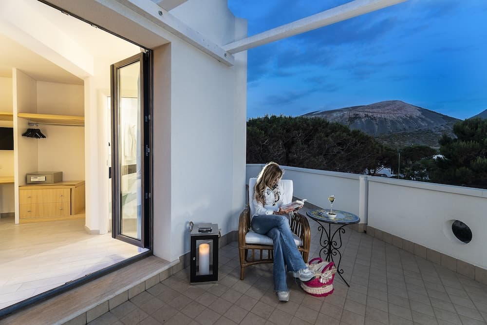 A serene evening unfolds on a cozy balcony overlooking lush greenery and distant hills, where a traveler enjoys a quiet moment with a book, glass of wine, and warm candlelight, ideal for a relaxing getaway.