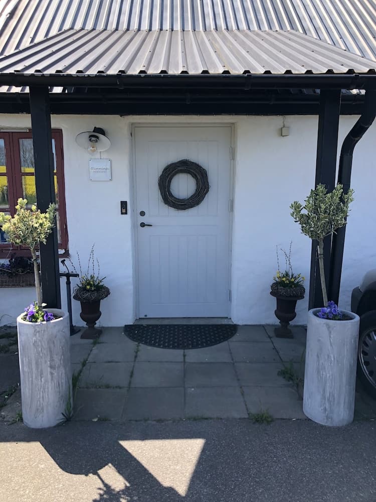 A charming white cottage entrance adorned with potted plants and a rustic wreath under a corrugated metal roof, evoking a cozy and welcoming atmosphere ideal for a serene getaway.