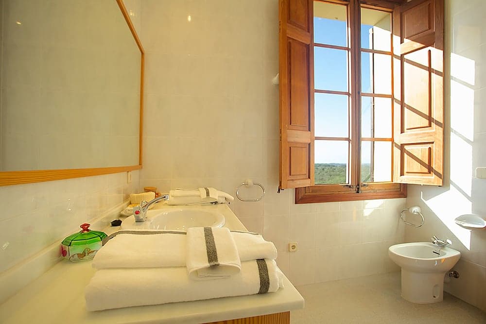 A serene bathroom with white tiled walls, neatly folded towels on the counter, a wooden-framed window offering a scenic view, and a bidet corner, ideal for travelers seeking comfort and tranquility in their accommodations.